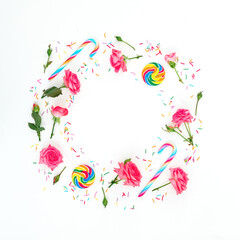 Round frame made of roses flowers with bright sugar candies on white background. Flat lay