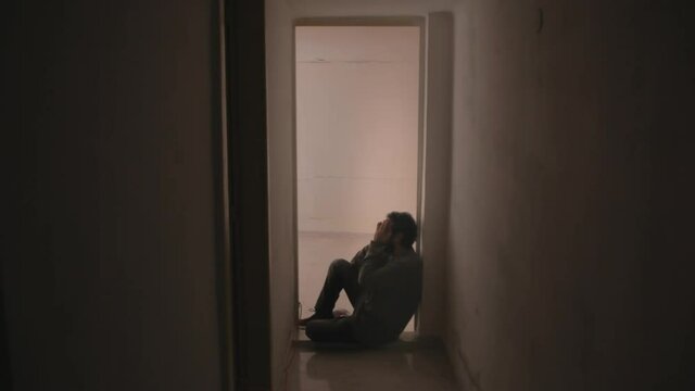A sad man, sitting on the floor near a door in his house, painfully crying in desperation, alone.
