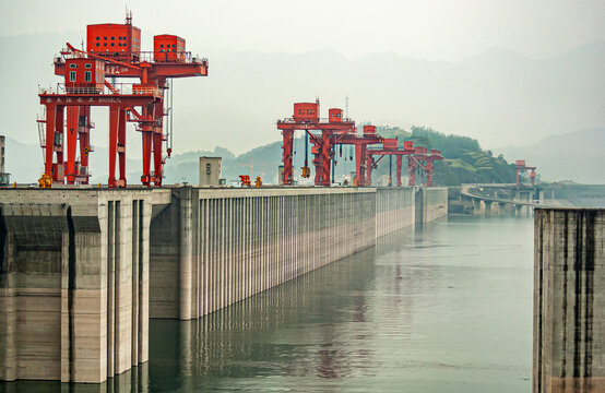 Three Gorges Dam, China - May 6, 2010: Yangtze River. Up-river shot of massive and long brown concrete dam with red flood control door lifts on top under foggy morning sky over hills and green water.