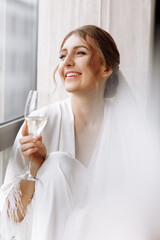 Bride s morning. Bride drinking champagne in the peignoir. young woman is sitting on a large window in a hotel room in bathroom. Beautiful girl in white wedding robe. wedding day