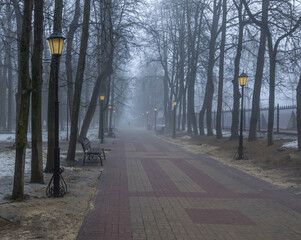 Misty morning park with deep fog, road leading far away in mist with trees and lamps on the sides