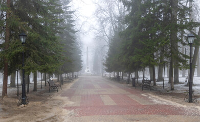 Misty morning park with deep fog, road leading far away in mist with trees and lamps on the sides