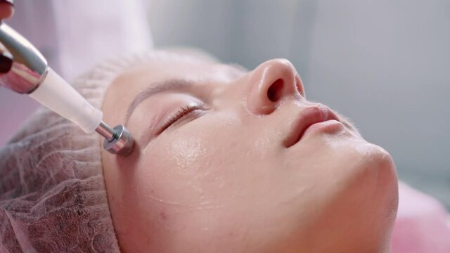 Young woman at the reception at the beautician. The doctor is doing microcurrent therapy of the facial skin. Side view. Detailed close-up.