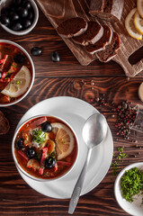Solyanka soup in a bowl with sausages, olives, tomatoes and pickled cucumber, on a dark background with lemons and bread
