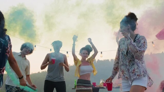 Low angle shot of young happy multiethnic friends dancing with colored smoke bombs at summer lake party