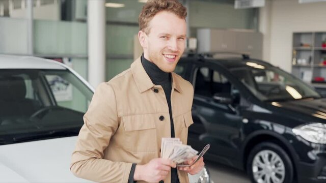 Happy young man dancing with money in hands on the background of new cars