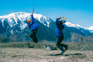 Two boys with toy weapons in their hands fight in a jump above the ground against the background of snowy mountains and blue clear sky