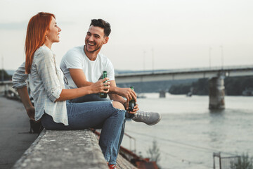 Young couple sitting on dock by the river ,relaxing and drinks beer.	
