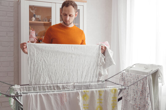 Man doing home chores. Caucasian man removes clothing and baby sheets after laundry from portable dryer in living room