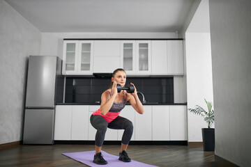 Fototapeta premium Side view of fit caucasian young woman squatting in kitchen. Strong girl wearing sportswear training legs on mat at home in morning carrying dumbbell in hands. Concept of home workout, weight loss.