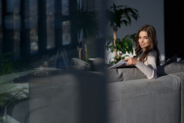 Side view of smiling young brunette beautiful woman sitting on sofa and working or resting with...