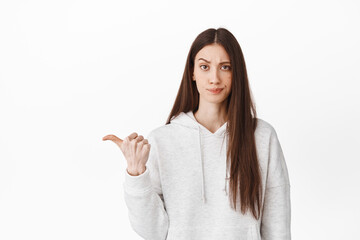 Doubtful and skeptical teenage girl frowning, looking with judging face and pointing aside at left logo banner, showing something bad or average, lame promo, white background