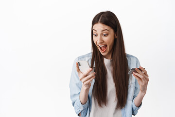 Online shopping. Image of girl winning money, screaming excited while looking at her phone, holding credit card, found super big discounts, standing over white background