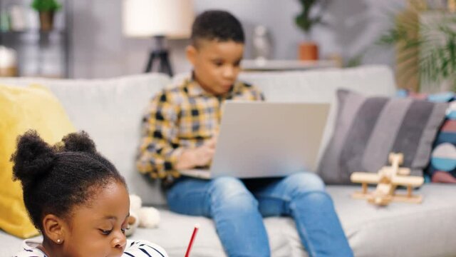Close up of little African American kids sitting in living room at home, cute girl drawing or writing on paper while her brother boy browsing online on laptop, e-learning education, children concept