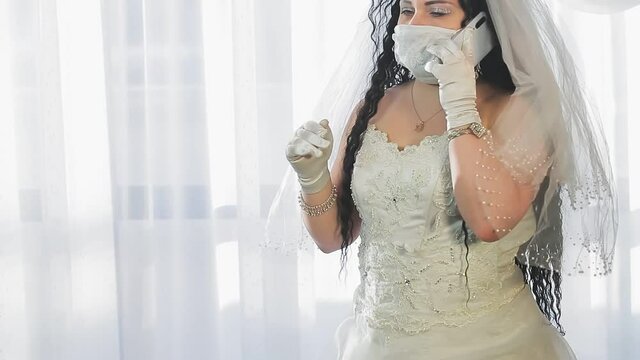 A Jewish bride in a wedding dress and a veil wearing a medical mask in the synagogue hall before the chupah ceremony speaks on a smartphone. Medium plan