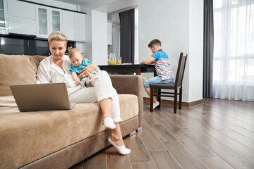 Attractive mother holding cute baby boy on hands while sitting on couch with laptop. Teenager studying near at kitchen table. Lifestyle routine of happy family.