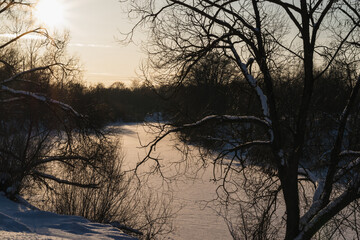 Snow-covered frozen winter river on a frosty winter day at sunset with shadows. View from the high bank through the branches of trees. Warm sunset sunlight. 