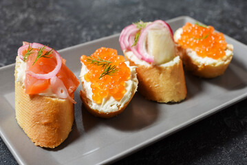 Bruschetta assortment with baguette, cream cheese, red caviar, salmon, herring and onion served on black stone background. Set of appetizers with various toppings