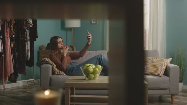 Beautiful girl in blue jeans and brown sweater lies on beige sofa on background of window, biting a red apple and and speaks via video link.