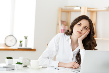 thoughtful woman with hand under chin bored at work, looking away sitting near laptop, demotivated...