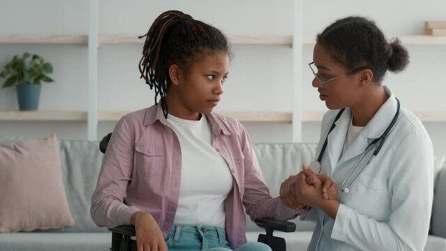 Doctor Examining Handicapped African Girl In Wheelchair Supporting Her Indoor