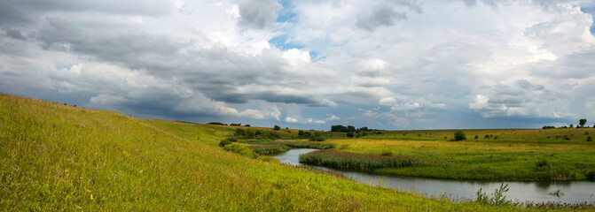 Summer panoramic landscape with dark stormy clouds over the calm river and green meadows.Russian rural landscape.
