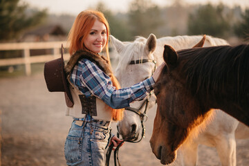 Cowgirl takes care of horses at ranch. Young red-haired woman affectionately stroking her horses and looking at camera.