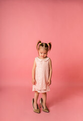 a guilty little girl in a pink party dress stands in her mother's shoes on a pink background with space for text