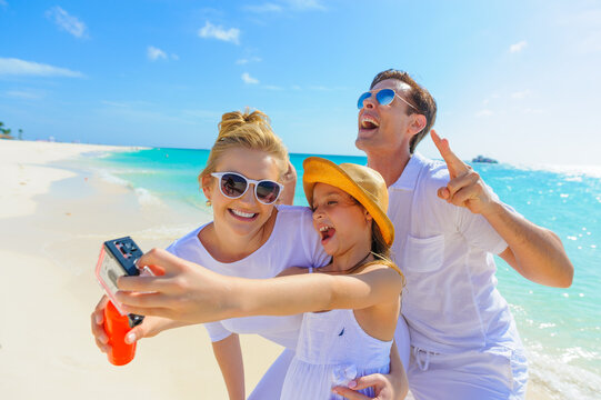 Family at the beach, mother, father and daughter taking photos, playing on the sand dressed in white tropical outfits