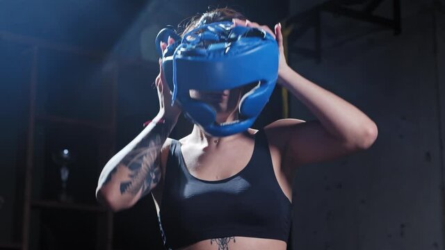 Tattooed woman puts on protective soft helmet and boxer gloves and stands in the fight pose