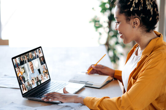 Inline briefing, conference. African American female manager communicates with business team during video call, listens financial webinar, takes notes, on laptop screen friendly multiracial people