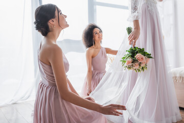 happy multicultural bridesmaids looking at bride holding wedding bouquet.