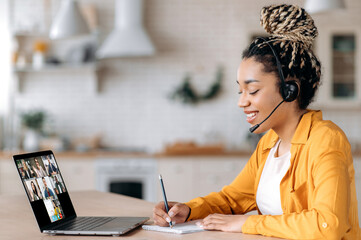 Happy African American female student listens to an online lecture using a laptop and a headset, takes notes, studies remotely, the teacher and other students are on the laptop screen. E-learning
