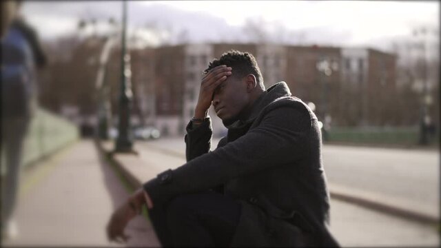 Worried black man sitting on floor in street having problems rubbing face with hand