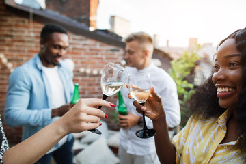 Close of two women toasting with glasses of champagne while two men drinking beer and chatting on background. Four mixed race friends enjoying party time on roof top.