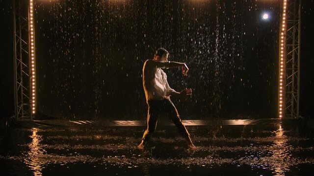 Young man sexy dancing in rain in unbuttoned white shirt. Wet clothing hugs muscular body. Raindrops glisten in studio light against black background. Lots of splashes and water droplets. Slow motion.