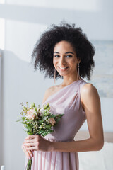 Elegant African American bridesmaid smiling at camera while holding wedding bouquet. - obrazy, fototapety, plakaty