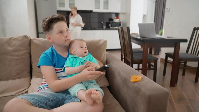 Teenage boy playing with his little brother on couch and watching cartoons on TV. Mother standing on background and having mobile conversation.