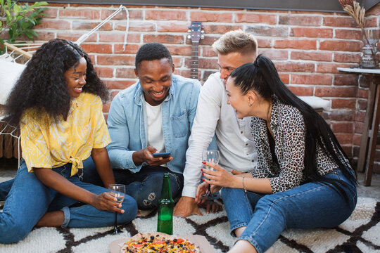 Group of four diverse people sitting together on open terrace and looking at mobile screen. Young men and women drinking alcohol, eating pizza and chatting during weekends.