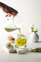 Women's hand pours green Matcha tea into the glass with ice in a white kitchen,with sweet macarons and ceramic vase on background.Close up of green tea.