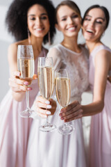 happy bride with interracial bridesmaids holding champagne glasses on blurred background.