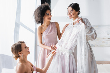 cheerful bride holding white wedding dress near multicultural bridesmaids.
