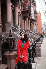 Fototapeta na wymiar a caucasian woman with dark hair and wearing a red coat stands outside in the city, on a sparsely populated street. a stylish woman traveling alone or with a friend and enjoying life, modern