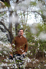 Photo of a young and attractive man in the park surrounded by almond trees during spring. He's wearing a jumper and smiling