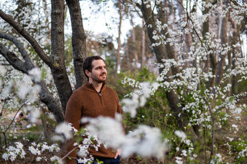 Photo of a young and attractive man in the park surrounded by almond trees during spring. He's wearing a jumper and smiling