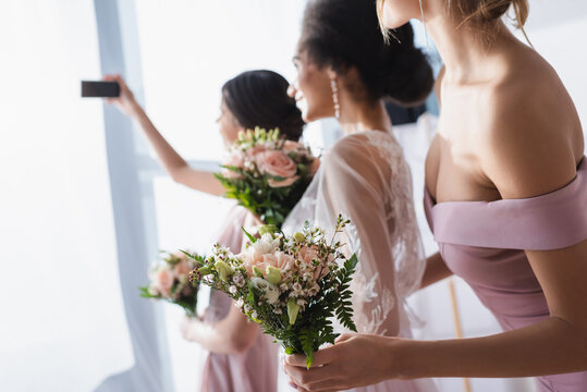 bride taking selfie with interracial bridesmaids holding wedding bouquets, blurred background.