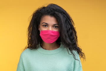 Close-up portrait of attractive pretty beautiful cute young dark-skinned woman wearing protecting pink facial mask isolated over bright colored orange yellow background