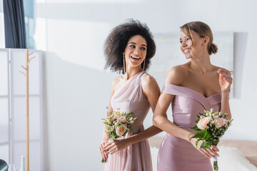 joyful multicultural bridesmaids holding wedding bouquets in bedroom. - obrazy, fototapety, plakaty