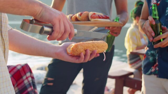 Tilt up shot of man taking bbq sausage from grill with metal tongues and putting it into bun while making hot dogs with help of friends on summer lake party