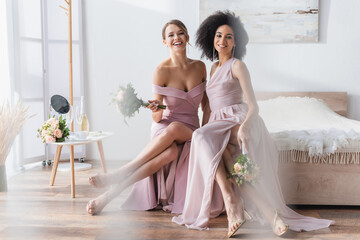 charming multicultural bridesmaids sitting on bed with wedding bouquets.